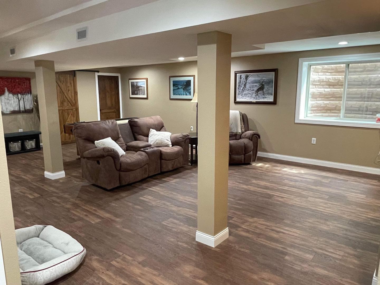 Finished basement with wood-look flooring, trimmed columns, seating, and a large window